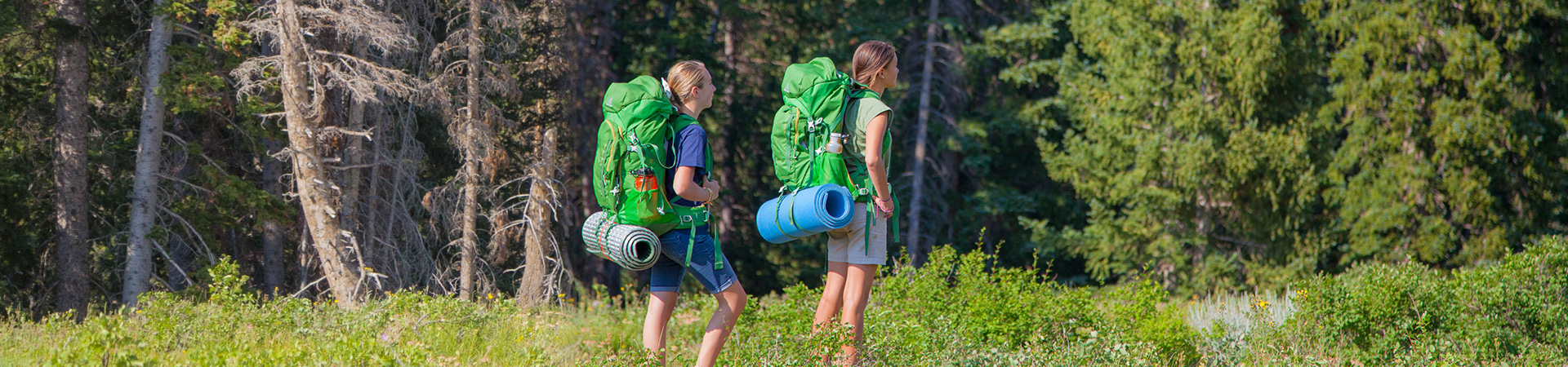  two girl scouts carrying large backpacks hiking through the woods 