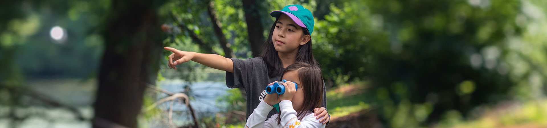  older girl scout points to the distance while younger girl scout looks through binoculars 
