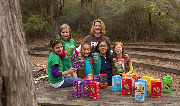 group of girl scouts and troop leader pose with boxes of cookies on a picnic table in woods